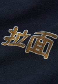 Tessuto nero con lettere in oro testurizzato. Il testo presenta un carattere bold e moderno con contorni ben definiti e una leggera lucentezza.