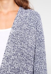 Pletený kardigan s modro-bílým texturovaným vzorem, s otevřeným předním designem a bez zapínání. Měkká a lehká tkanina.