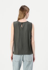 Ermeløs, plisséert topp i mørkegrønn stoff; har en nøkkelhulls baklukking og en jevn tekstur. Kombinert med lyseblå jeans.