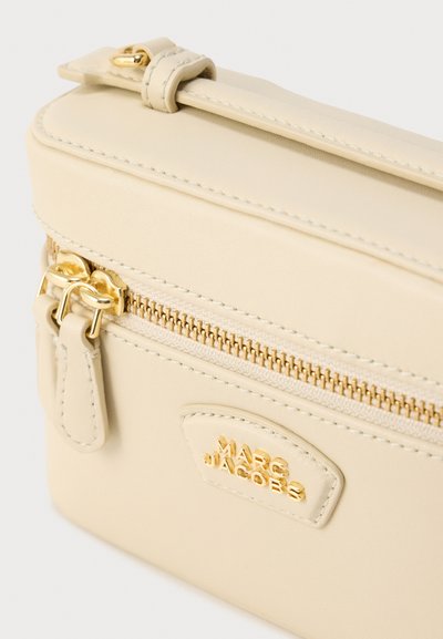 Τσάντα από δέρμα ανοιχτού μπεζ χρώματος με υφή και χρυσούς μεταλλικούς μηχανισμούς· διαθέτει φερμουάρ, μικρή λαβή και ανάγλυφο λογότυπο.