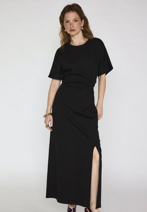 Donna con capelli ondulati che indossa un vestito lungo nero a maniche corte con uno spacco laterale, accessoriata con orecchini a cerchio dorati e braccialetti.