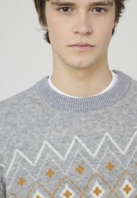 Pull tricot gris avec un motif géométrique aux accents blancs et dorés. Le col côtelé apporte de la texture au design décontracté.