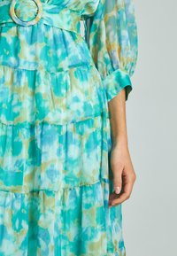 Abito a strati in stampa acquerello blu chiaro, verde e beige. Presenta una ampia cintura con una fibbia circolare e maniche lunghe a sbuffo.