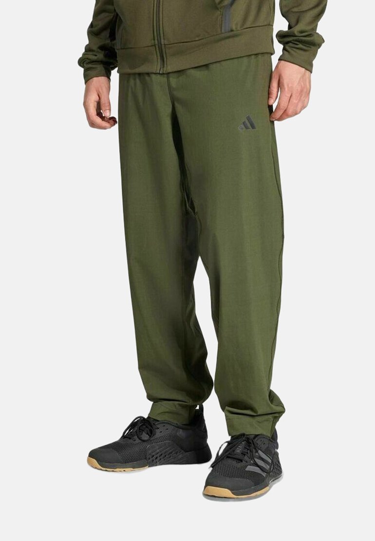 Homme portant un pantalon de survêtement Adidas vert olive et des chaussures de sport noires, debout devant un fond uni.