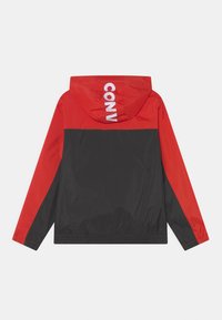 Rød og sort vindjakke med hætte. Har stort hvidt "CONV" tekst på ryggen, elastisk linning og glat, shiny stof.