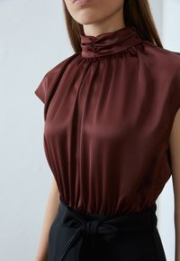 Bordeaux satijnen blouse met een geschulpte hoge hals, korte mouwen en geplooide details; gecombineerd met een op maat gemaakte zwarte tailleband.