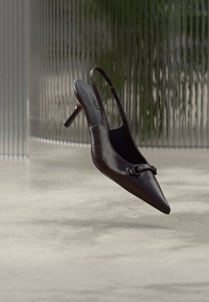 Escarpin slingback noir brillant à bout pointu avec une petite boucle décorative, flottant au-dessus d'un sol en béton avec un fond en verre texturé.