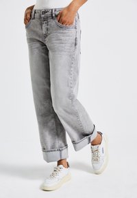 Persona vestita con jeans grigi chiari con risvolti e tasche frontali, in piedi con le mani nelle tasche.