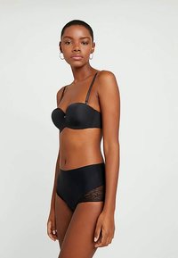 Mulher a modelar um soutien preto sem alças e umas cuecas altas coordenadas com painéis de renda, em pé contra um fundo claro e liso.