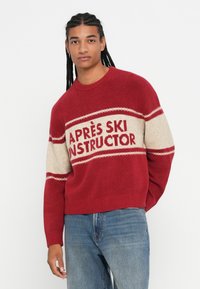 Fiatal férfi hosszú fonott hajjal, piros és bézs pulóvert viselve, amelyen az "APRÈS SKI INSTRUCTOR" felirat szerepel, kék farmerben, fehér háttér előtt pózol.