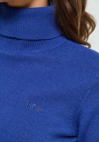 Pull à col roulé bleu en maille côtelée. Présente un petit logo brodé dans une couleur assortie sur le devant.
