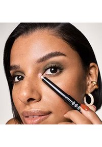 Matita eyeliner nera tenuta da una persona con capelli scuri e tonalità di pelle neutra, che mostra l'applicazione sull'occhio interno. Ombretto verde tenue applicato.