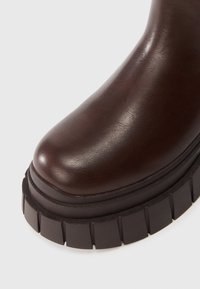 Hnědé syntetické kožené kotníkové boty s kulatou špičkou a silnou, strukturovanou černou podrážkou s výraznými drážkami. Minimalistický design, detailní pohled.