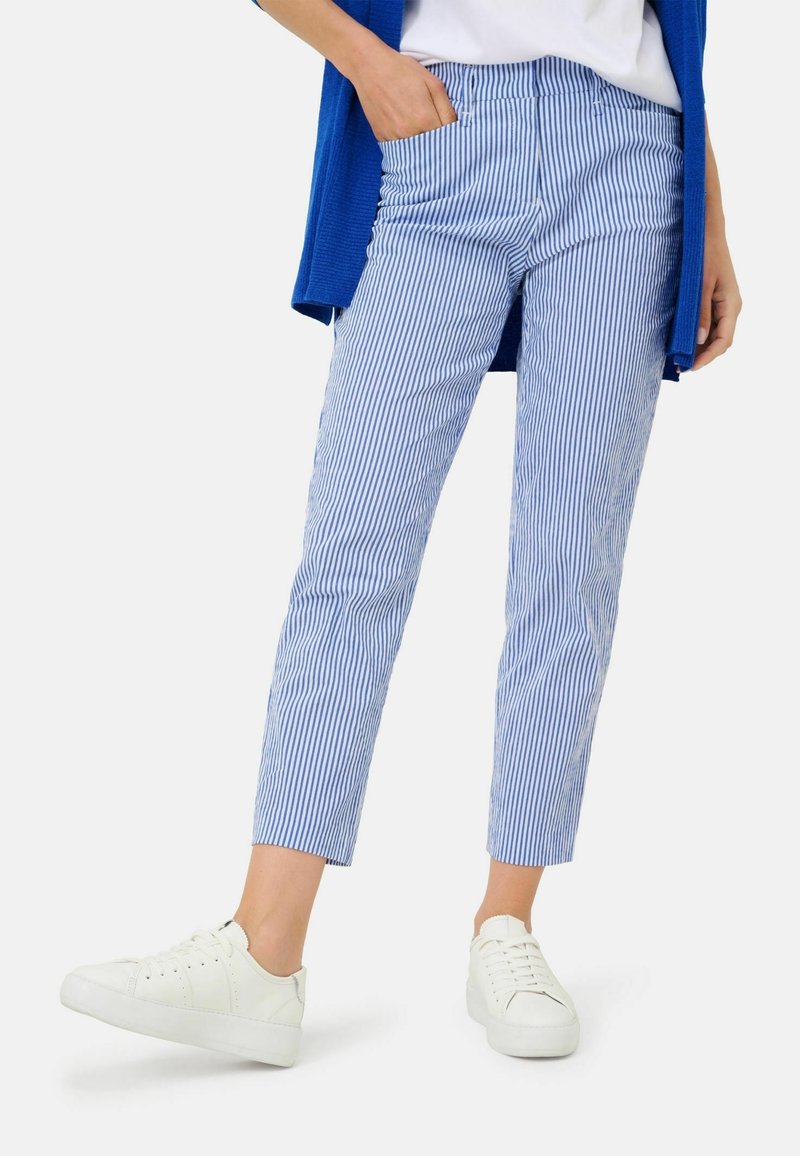 Personne portant un pantalon court à rayures verticales bleues et blanches, des baskets blanches, un haut blanc et un cardigan bleu, debout avec une main dans la poche.
