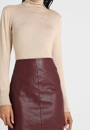 Femme portant un pull à col roulé beige et une jupe en cuir rouge foncé taille haute, vue du cou jusqu'à la mi-cuisse.
