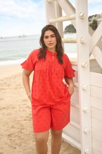 Femme aux longs cheveux foncés portant un chemisier rouge à motifs à manches courtes et un short assorti, debout sur une plage, appuyée contre une structure en bois blanche.