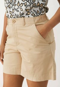 Shorts en coton beige avec poches latérales, taille régulière et ourlet cousu. Portés avec un haut à motifs présentant un design floral noir et jaune.