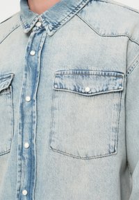 Ανοιχτό μπλε πουκάμισο τζιν με κλασικό γιακά, δύο μπροστινές τσέπες με κουμπωτές πατιλέτες και ασημί κουμπιά κατά μήκος της λωρίδας κουμπώματος.