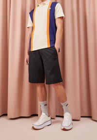 Polo beige, marine et orange avec des rayures verticales, associé à un short noir, des chaussettes grises et des baskets blanches avec des semelles marron.