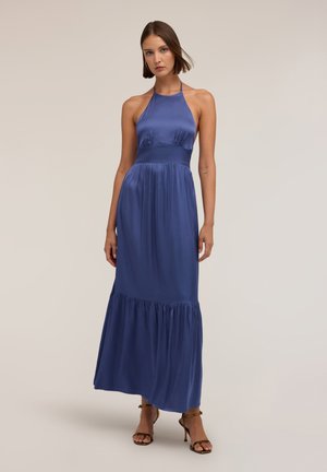 Frau trägt ein ärmelloses blaues Satin-Maxikleid mit Neckholder und stufigem Rock sowie braune Sandalen mit hohen Absätzen, steht vor einem schlichten Hintergrund.