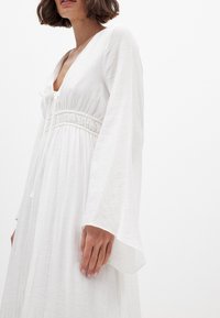 Hvid langærmet kjole med dyb V-hals, smocket talje og flydende tekstur. Har letvægtsstof med let krøllet detalje.