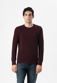 Pull en maille bordeaux avec un col rond, des poignets et un ourlet côtelés, arborant un petit logo sur la poitrine. Associé à un jean bleu.