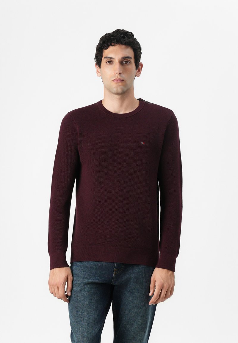 Pull en maille bordeaux avec un col rond, des poignets et un ourlet côtelés, arborant un petit logo sur la poitrine. Associé à un jean bleu.