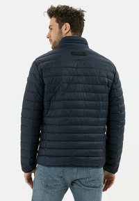 Dunkelblaue Pufferjacke mit hohem Kragen, gestepptem Design und zwei schwarzen Druckknöpfen auf der Rückseite. Trägt man mit hellblauen Jeans.