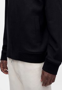 Homme portant une veste noire zippée avec poches latérales et poignets côtelés, associée à un pantalon écru, visible du milieu du torse jusqu'aux mains.
