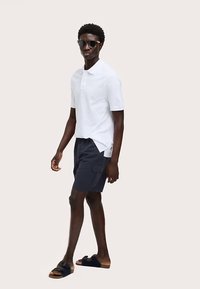 Bílá polokošile, námořnicky modré cargo šortky, černé sandály a sluneční brýle. Model stojí bokem na neutrálním pozadí.