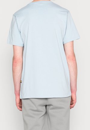 Světle modré tričko s krátkými rukávy a kulatým výstřihem, hladká textura a malá logo etiketa na boku. Nošené s šedými tepláky.