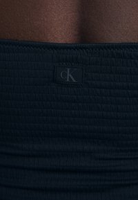 Čierna priliehavá tkanina s textúrovaným vzorom a logom s názvom "cK" vyšitým na páse. Hladký a elastický materiál.