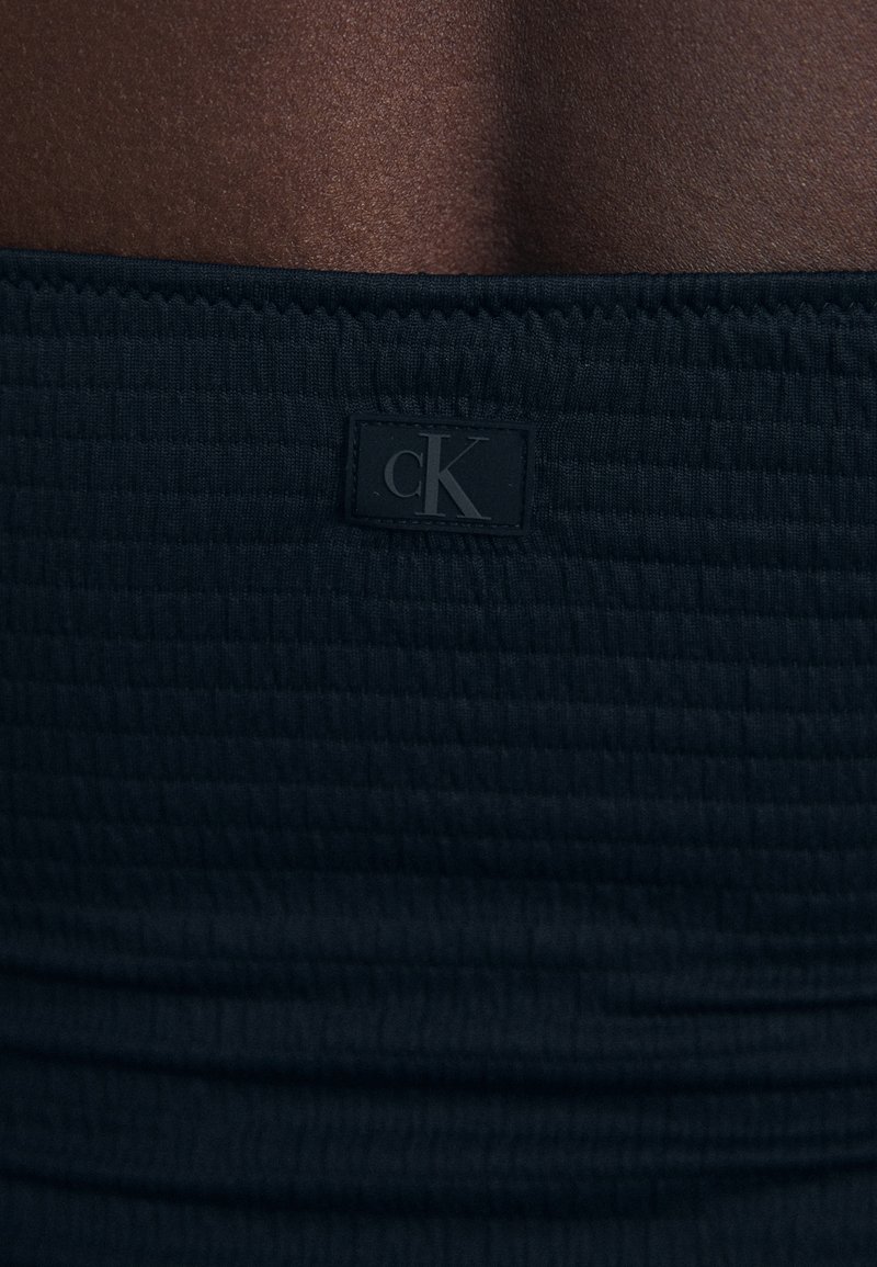 Czarna dopasowana tkanina z teksturowanym wzorem i naszytą metką z logo "cK" na pasku. Gładki i elastyczny materiał.
