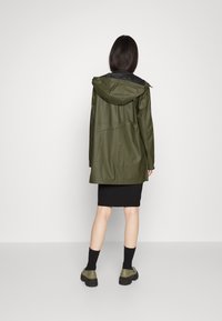 Giacca impermeabile verde oliva con cappuccio, maniche lunghe e vestibilità morbida, indossata sopra una gonna nera a coste e abbinata a scarpe verdi robuste.