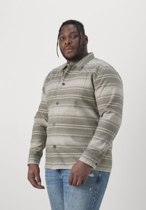 Olivgrünes Hemd mit Knopfleiste, horizontalen Streifen, strukturiertem Stoff, langen Ärmeln und einer Brusttasche; getragen mit blauen, verwaschenen Jeans.