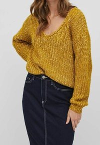 Senfgelbes Strickpullover mit V-Ausschnitt und langen Ärmeln, kombiniert mit einem dunklen Jeans-Bleistiftrock mit weißen Nähten.