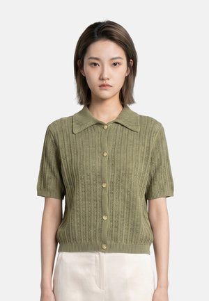 Giovane donna con capelli corti e scuri che indossa un cardigan verde lavorato a maglia con maniche corte e bottoni e pantaloni chiari su uno sfondo neutro.