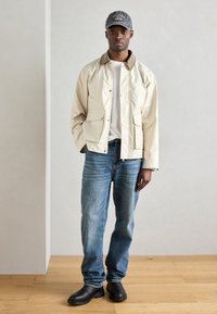 Μπεζ βαμβακερό μπουφάν με καφέ γιακά, δύο μπροστινές τσέπες, συνδυασμένο με μπλε τζιν και μαύρες παντόφλες. Γκρίζο καπέλο με κείμενο από πάνω.