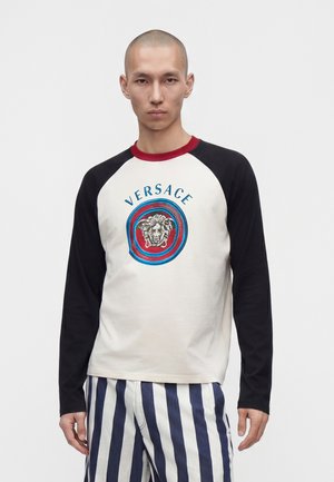 Homme portant une chemise blanche à manches longues avec des manches noires et un col rouge, ornée d'un logo Méduse et du texte « Versace », associée à un pantalon bleu et blanc à rayures.