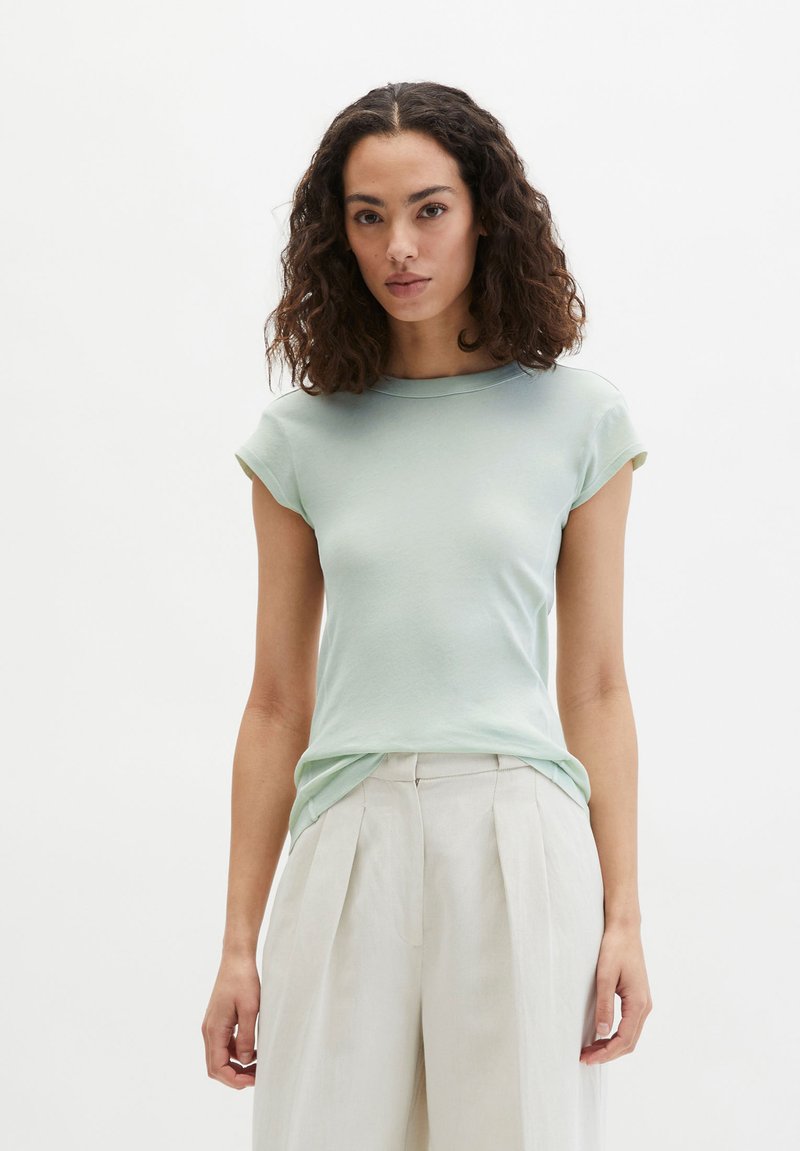 Frau mit lockigem dunklem Haar, die ein hellgrünes kurzärmeliges Oberteil trägt, das in eine hoch taillierte, beige plissierte Hose gesteckt ist, stehend vor einem weißen Hintergrund.