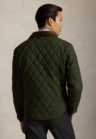 Hombre de espaldas, vistiendo una chaqueta acolchada verde oscuro con cuello marrón y pantalones grises sobre un fondo beige liso.