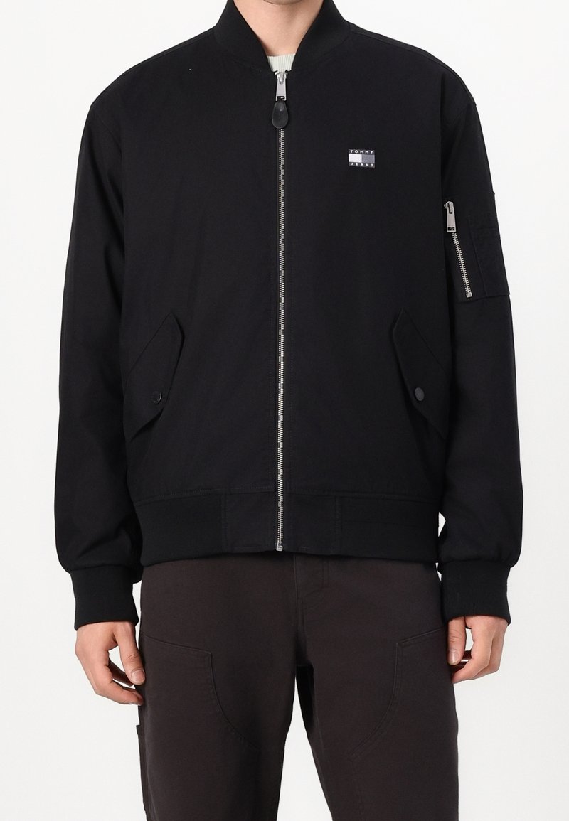 Homme portant une veste bomber noire avec fermeture éclair devant, poches à rabat, un petit logo sur la poitrine et une poche zippée sur la manche gauche.