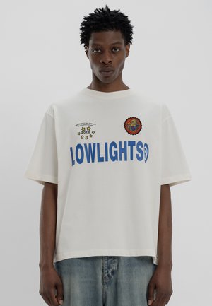 T-shirt oversize in cotone color crema con testo blu "LOWLIGHTS", toppe ricamate e girocollo. Vestibilità casual, maniche corte.