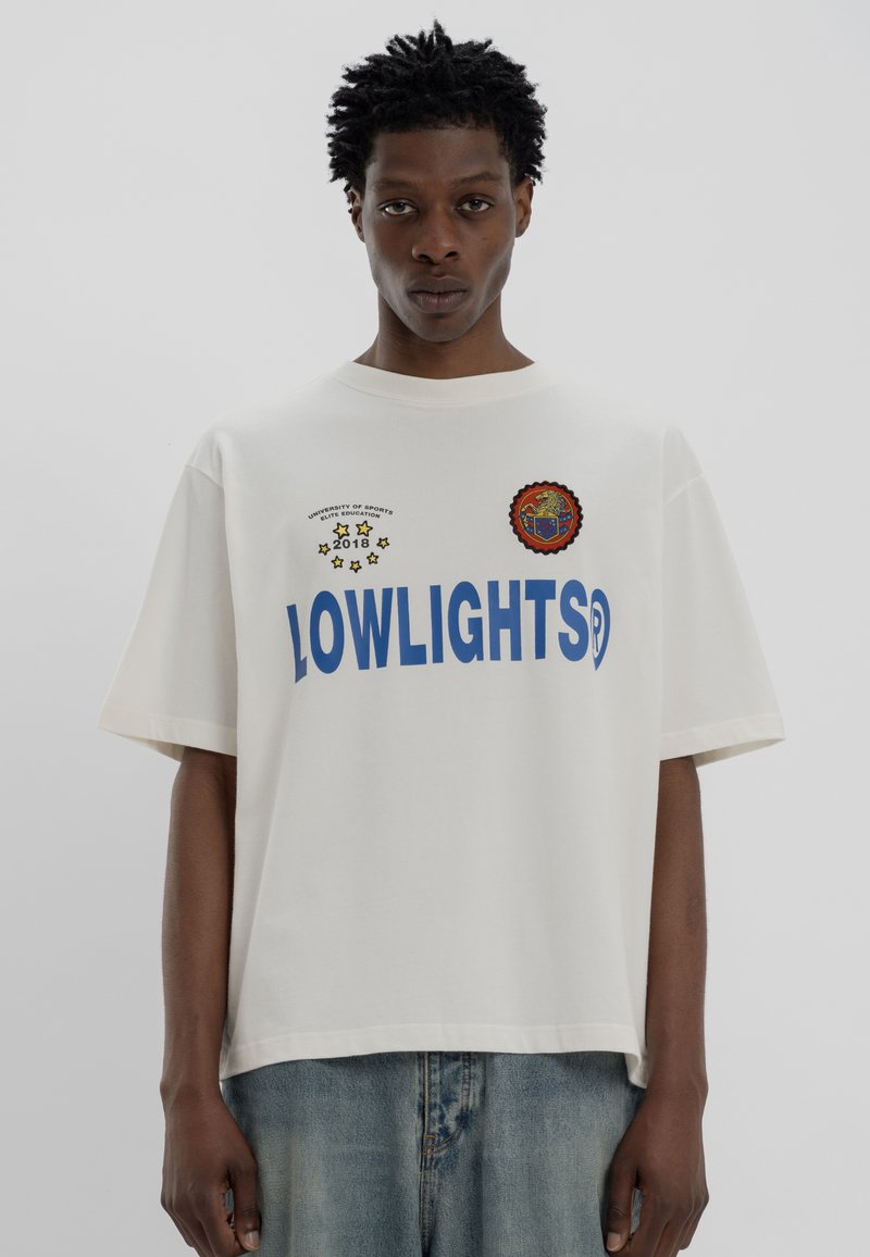 T-shirt en coton crème surdimensionné avec un texte bleu "LOWLIGHTS", des patchs brodés et un col rond. Coupe décontractée, manches courtes.