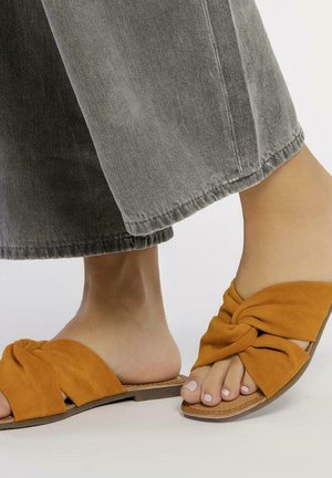 Infradito in suede marrone con design annodato, suole piatte e punte aperte, abbinate a pantaloni larghi grigi. Texture liscia e stile casual.