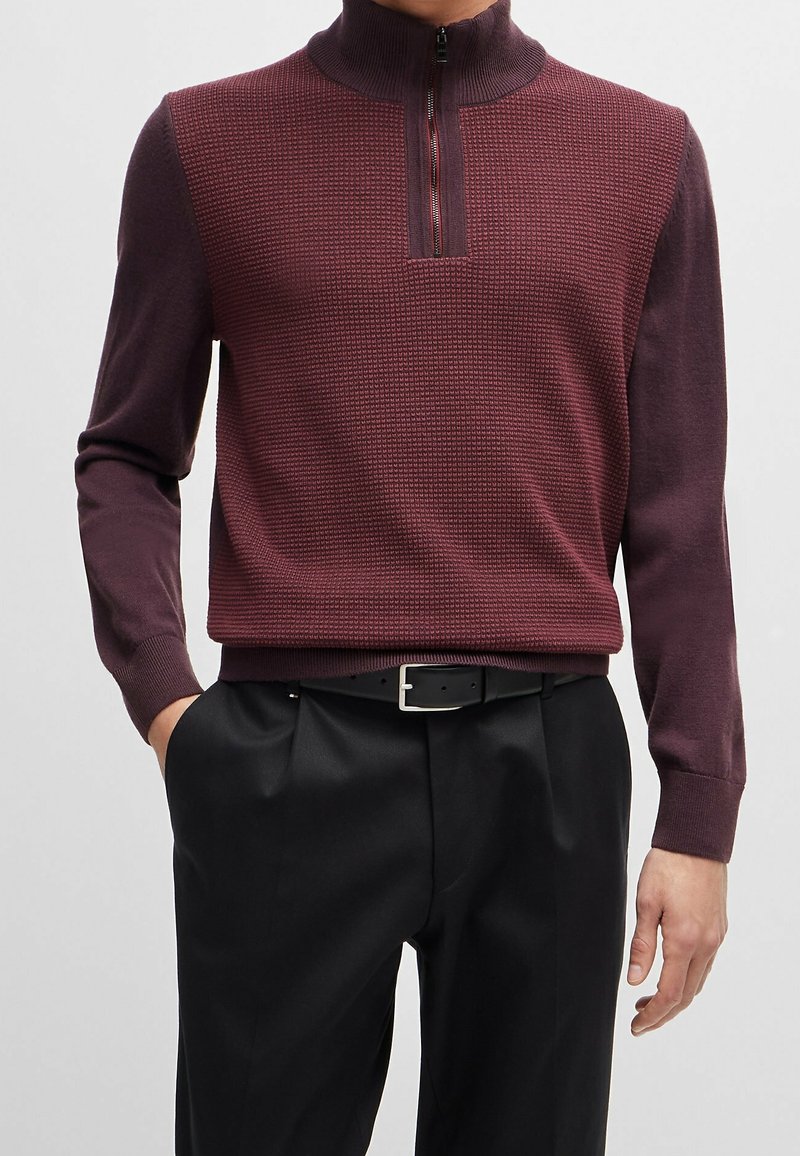 Burgundy half-zip trui met een gestructureerd patroon en geribbelde kraag. Lange mouwen en een aansluitend ontwerp, gecombineerd met zwarte broeken en een riem.