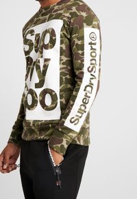 Sudadera con patrón de camuflaje en verde y marrón, con un cuadro de logo blanco que lleva el texto "Superdry" en el frente y una raya blanca en la manga.