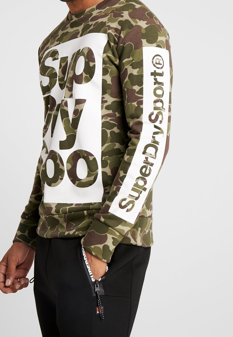 Sudadera con patrón de camuflaje en verde y marrón, con un cuadro de logo blanco que lleva el texto "Superdry" en el frente y una raya blanca en la manga.