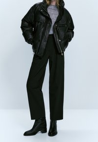 Personne portant une veste en cuir matelassée noire, une chemise grise, un pantalon large noir et des bottines noires, debout devant un fond clair uni.