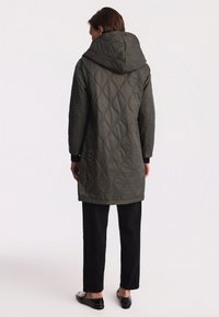 Pikowany oliwkowo-zielony długi płaszcz z kapturem, z teksturowanym falistym wzorem oraz czarnymi akcentami na rękawach, w połączeniu z czarnymi spodniami.